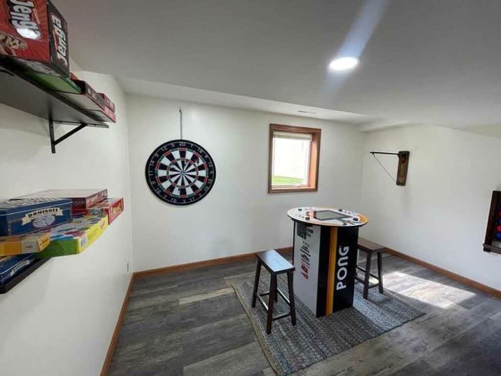 A game room with a dartboard on the wall, a round Pong table with two stools, and shelves holding various board games.