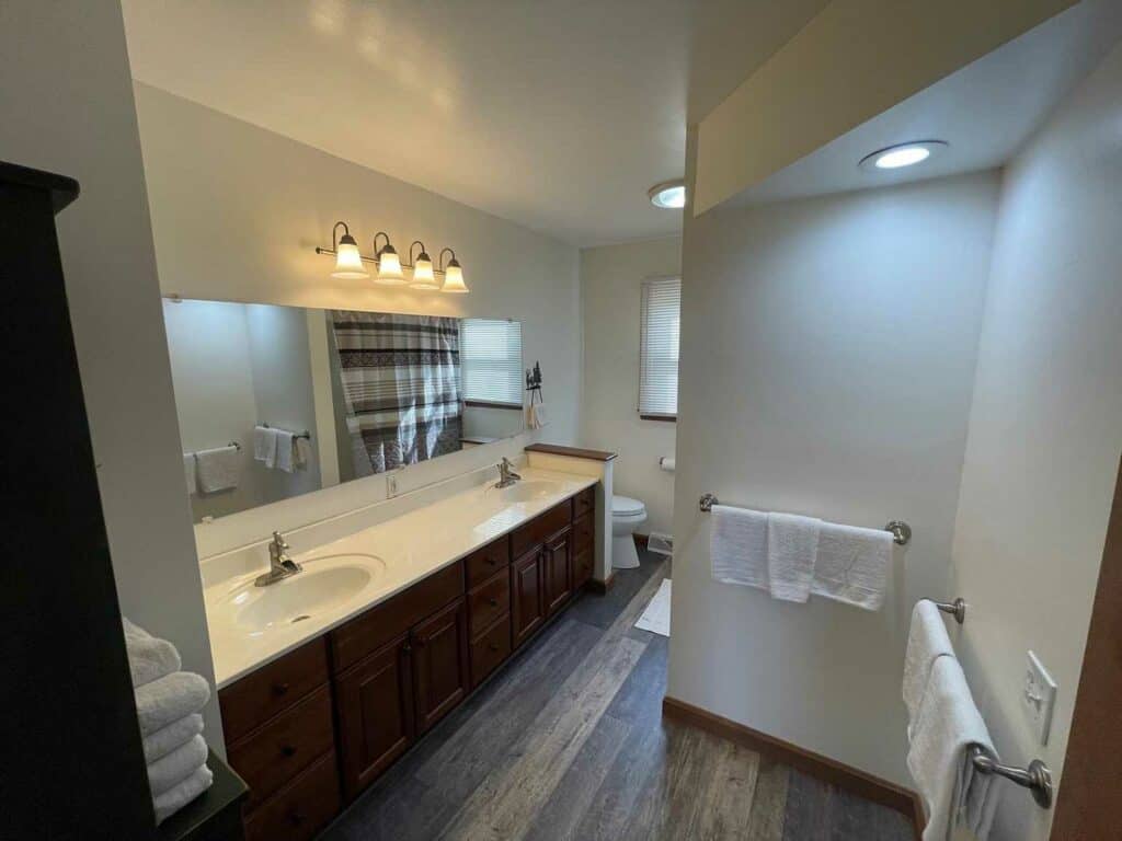 Spacious bathroom with double sinks, large mirror, wood cabinets reminiscent of cozy cabins, overhead lighting, a toilet, towel racks, and a window with blinds.