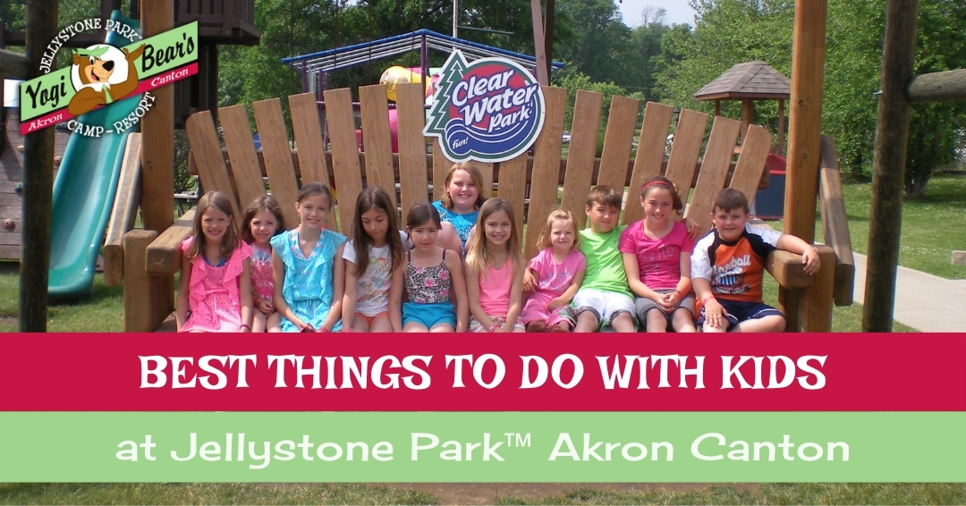 A group of children sit on a large wooden bench at Jellystone Park Akron Canton's Clear Water Park, with playground equipment visible in the background.