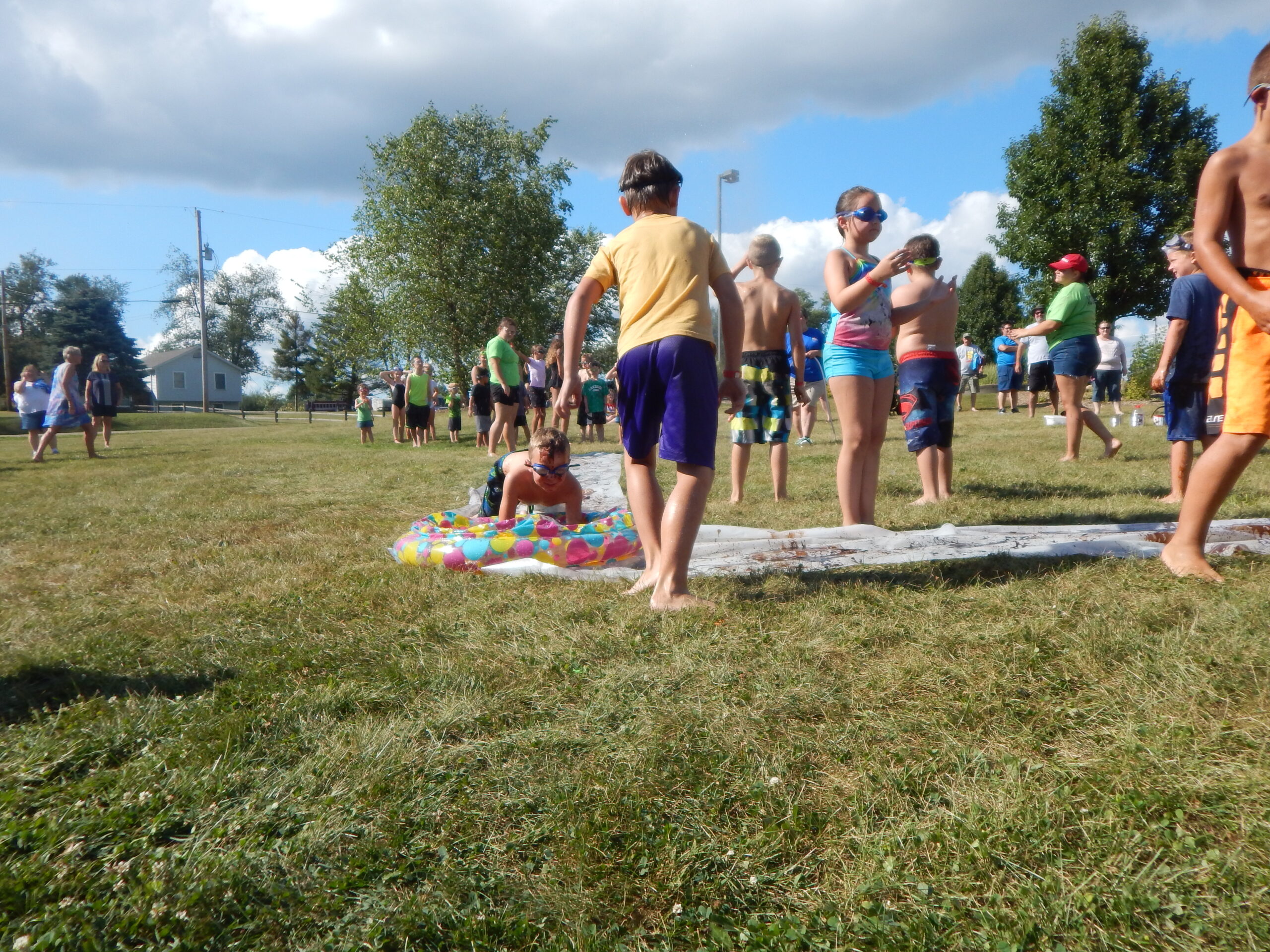 Children play outside on a grassy field at a family camping Ohio site, some standing on a slip-and-slide while others wait nearby; adults and more kids gather in the background under a mostly sunny sky.