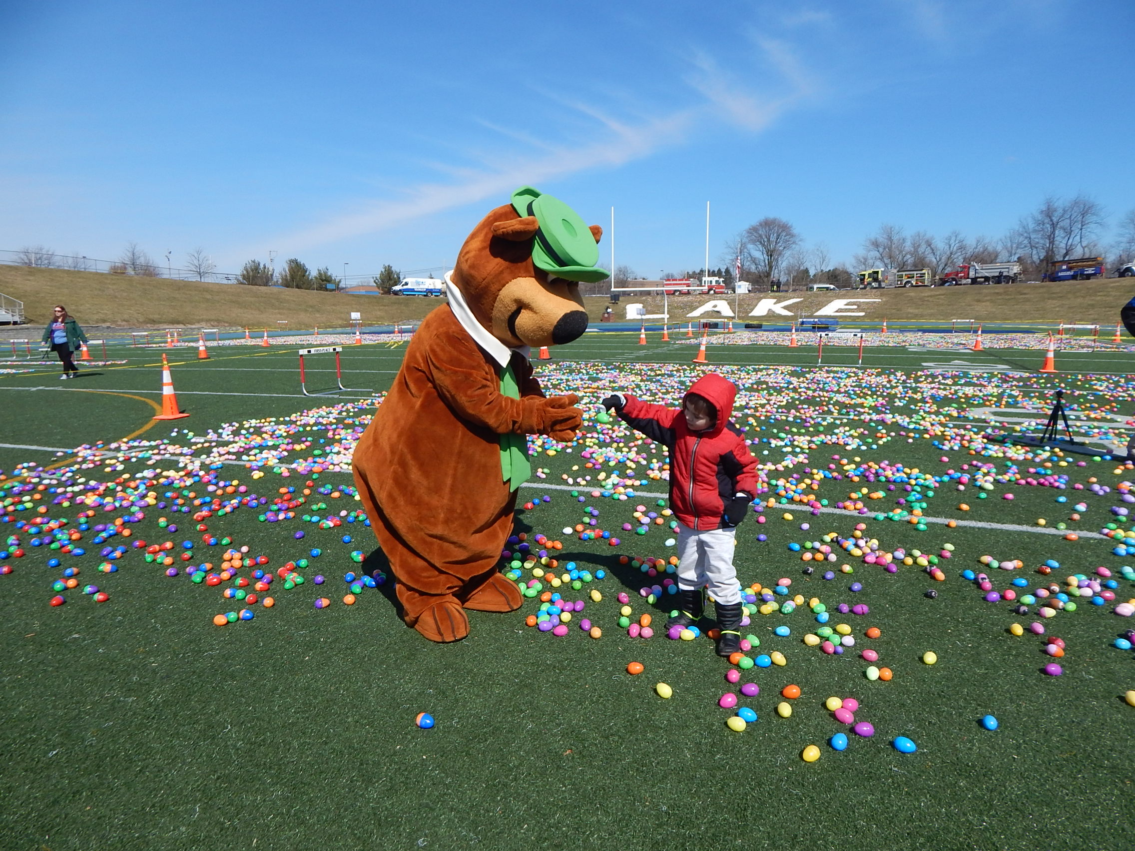 A person in a bear mascot costume shakes hands with a child on a field covered in colorful plastic eggs during an outdoor event. A person in a bear mascot costume shakes hands with a child on a field covered in colorful plastic eggs during an outdoor event.