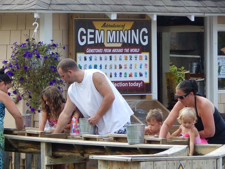 Adults and children participate in a gem mining activity, using sifters and buckets at an outdoor station, with a "Gem Mining" sign in the background.