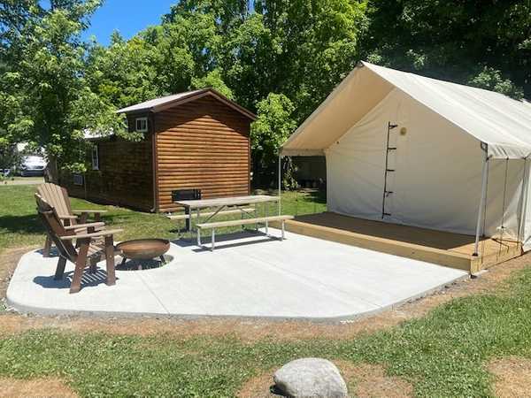 A white canvas tent and a small wooden cabin sit on a concrete pad with two wooden chairs, a picnic table, and a fire pit, surrounded by grass and trees.