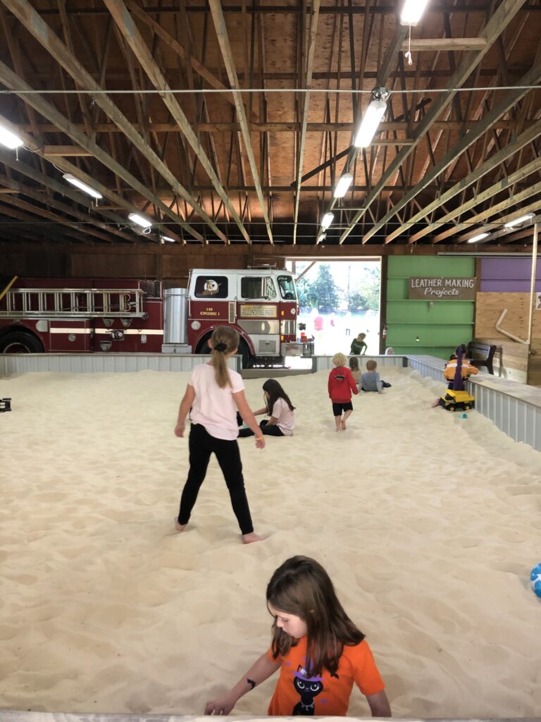 Children play in an indoor sand area, with a vintage fire truck and leather making sign visible in the background under a wooden roof.