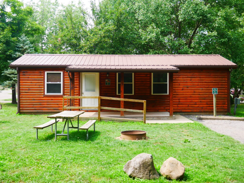 A small wooden cabin with a front porch and ramp, surrounded by green grass and trees, with a picnic table and fire ring in the foreground.