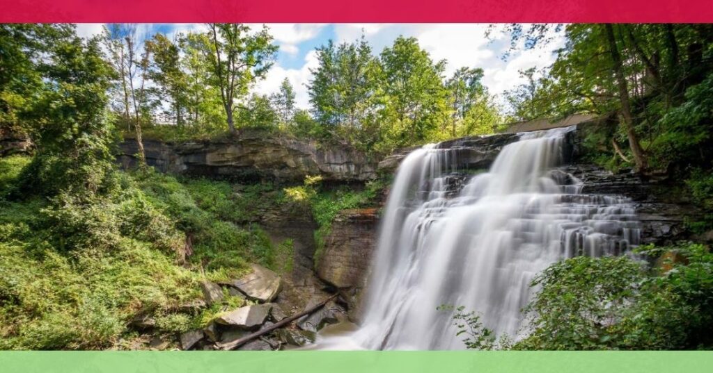 A wide waterfall cascades over a rocky cliff surrounded by lush green trees under a partly cloudy sky. There are red and green borders at the top and bottom of the image.