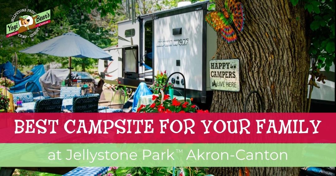 A family campsite at Jellystone Park Akron-Canton with a camper, picnic tables, umbrella, chairs, and a "Happy Campers Live Here" sign.