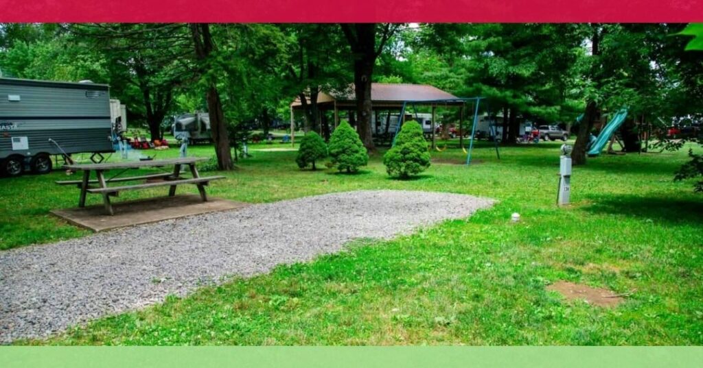 A gravel campsite with a picnic table, surrounded by grass and trees, with a playground, camper, and pavilion visible in the background.