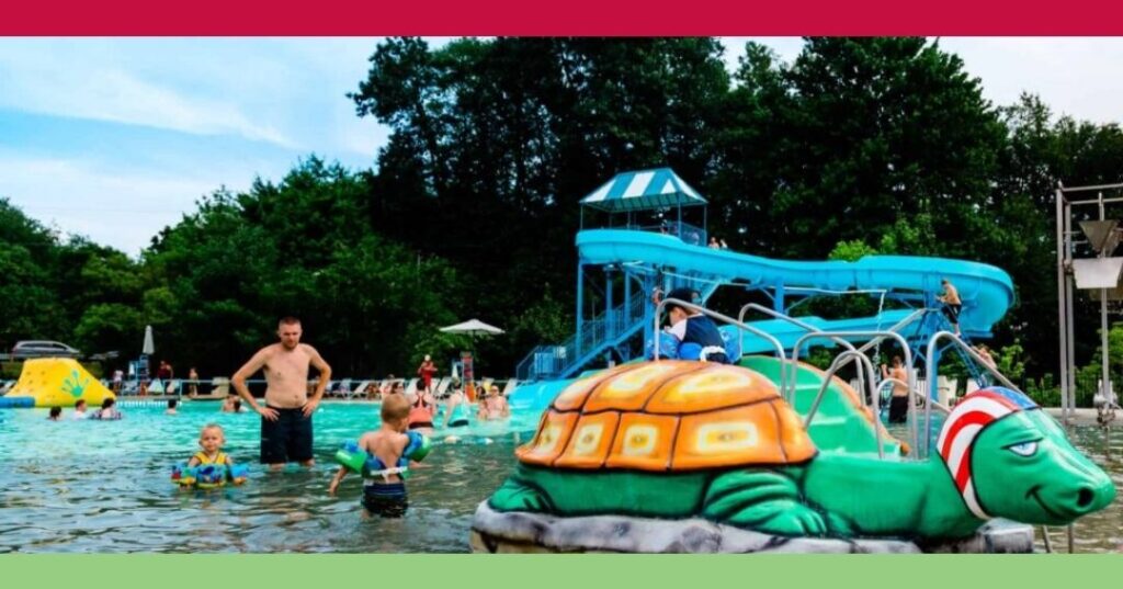 Children and adults play in an outdoor pool with a large green turtle structure and a blue water slide in the background. Trees and lounge chairs surround the pool area.