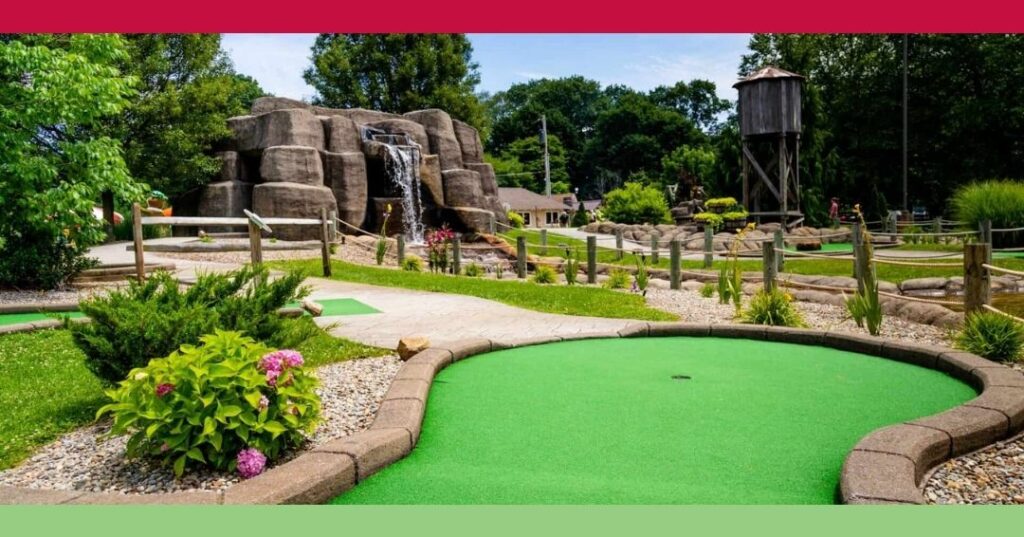 A colorful mini-golf course features artificial turf, landscaping, a rock waterfall, and a wooden water tower on a sunny day.