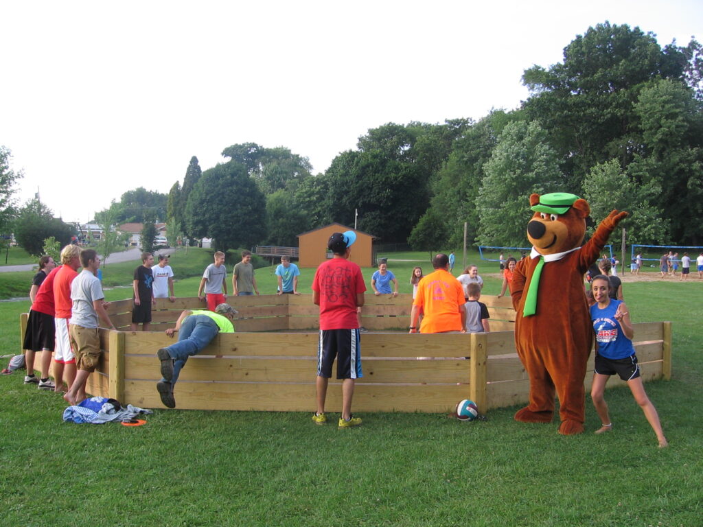 A group of people play gaga ball outdoors while a person in a bear costume waves and poses for a photo next to a smiling girl.