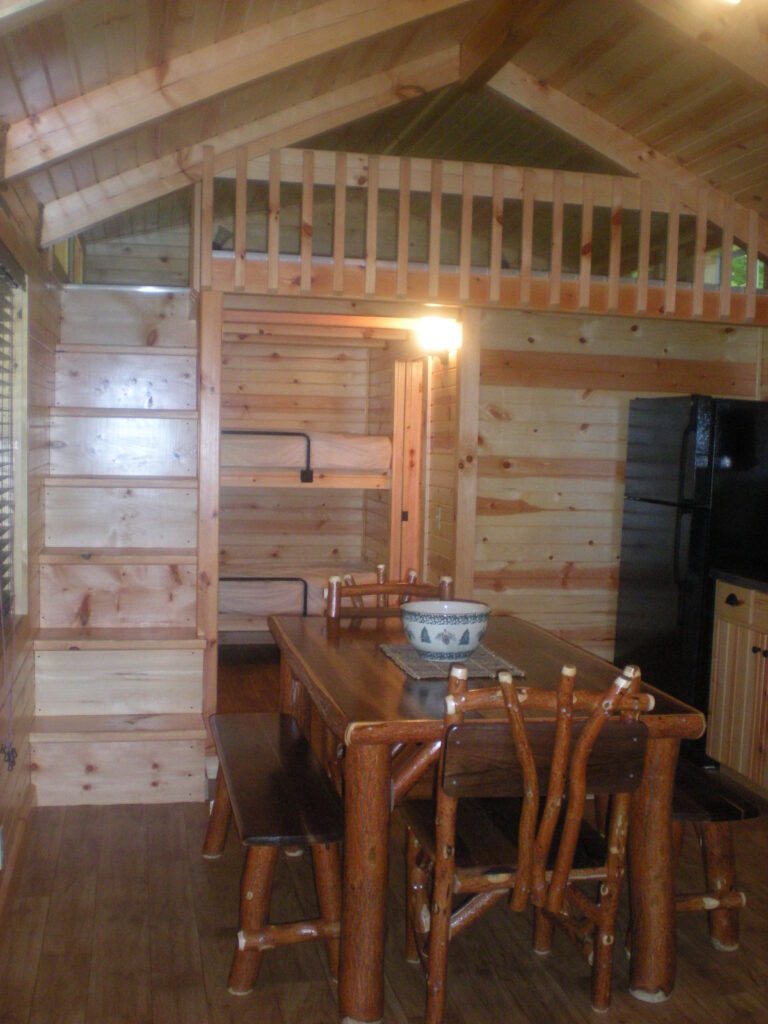 Interior of a wooden cabin with a dining table and benches, a loft area with a railing above, and a black refrigerator in the kitchen area.