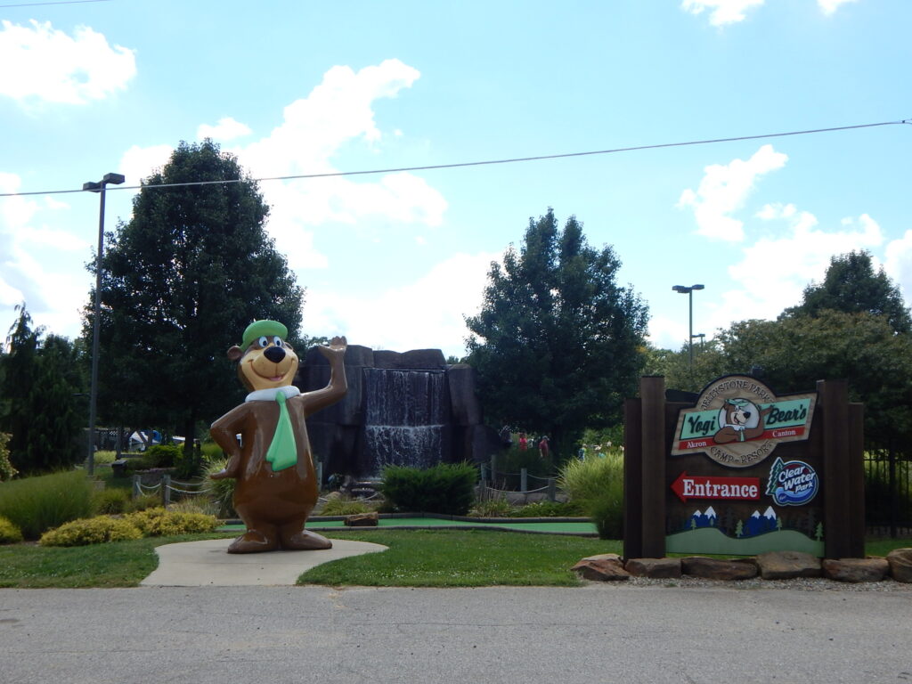 A large Yogi Bear statue stands near the entrance sign for Yogi Bear’s Jellystone Park, with trees and a waterfall in the background.