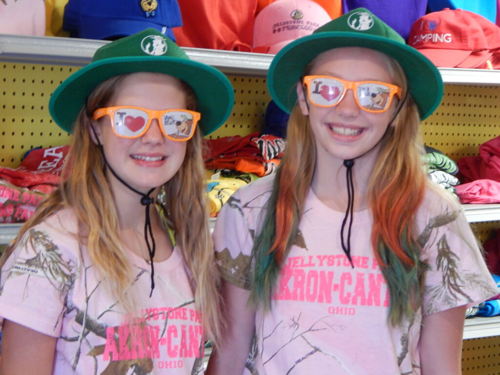 Two girls wearing matching green hats, orange sunglasses, and pink Yellowstone Park T-shirts stand in front of a display of colorful hats and shirts.