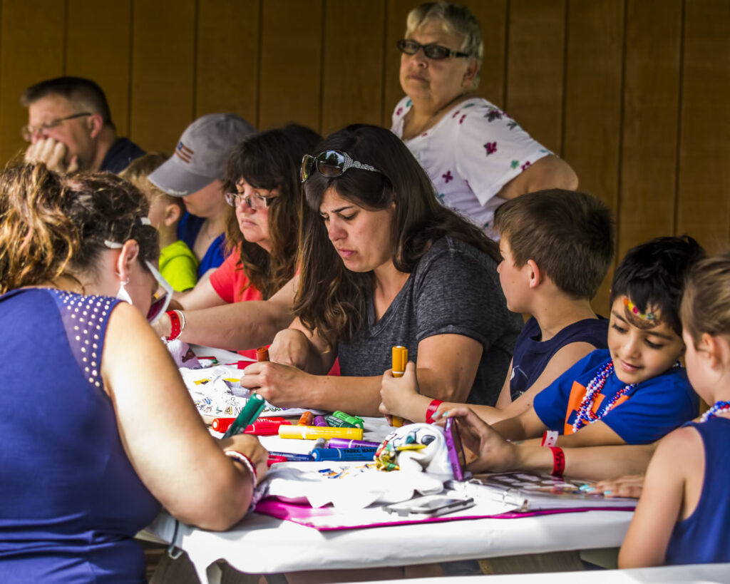Adults and children sit around a table engaged in arts and crafts activities, with various supplies spread out; a few adults observe in the background.
