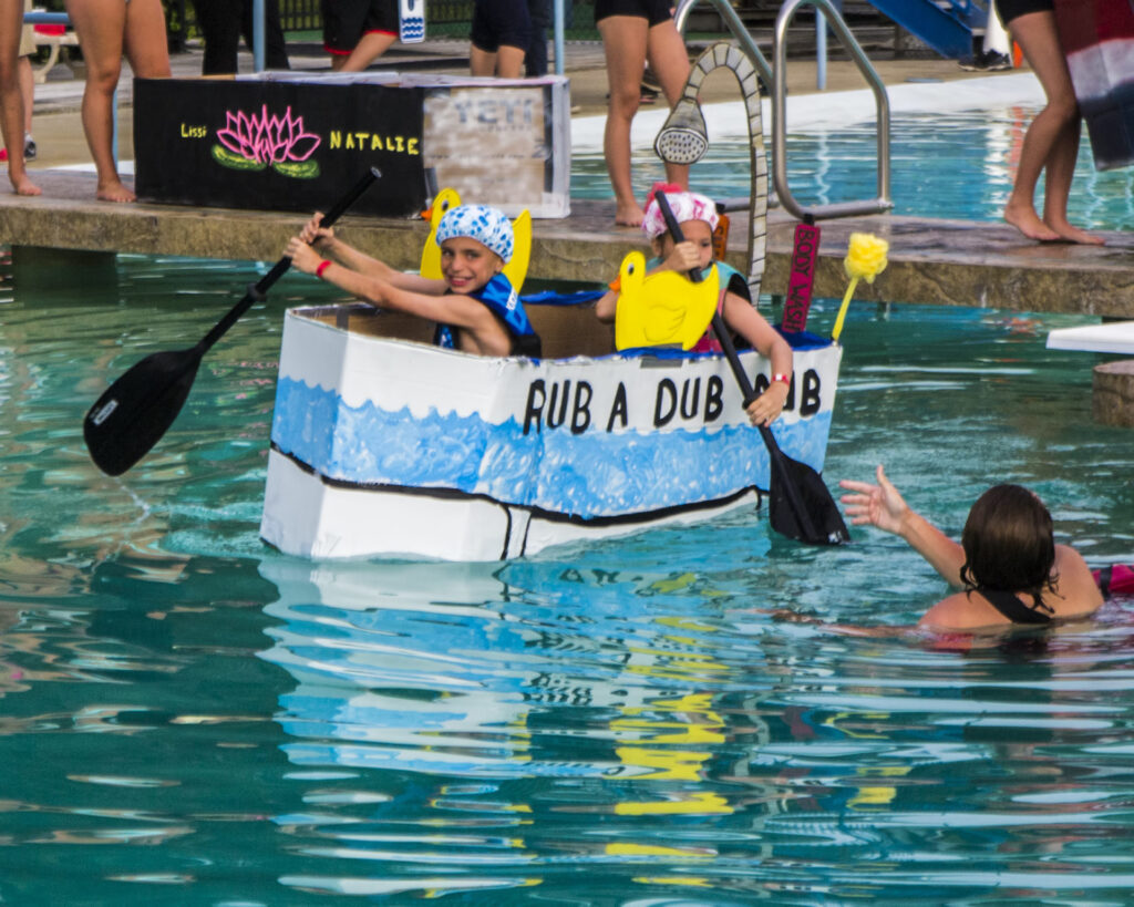 Two children paddle a homemade cardboard "bathtub" boat labeled "RUB A DUB DUB" in a pool while others watch from the water and poolside.