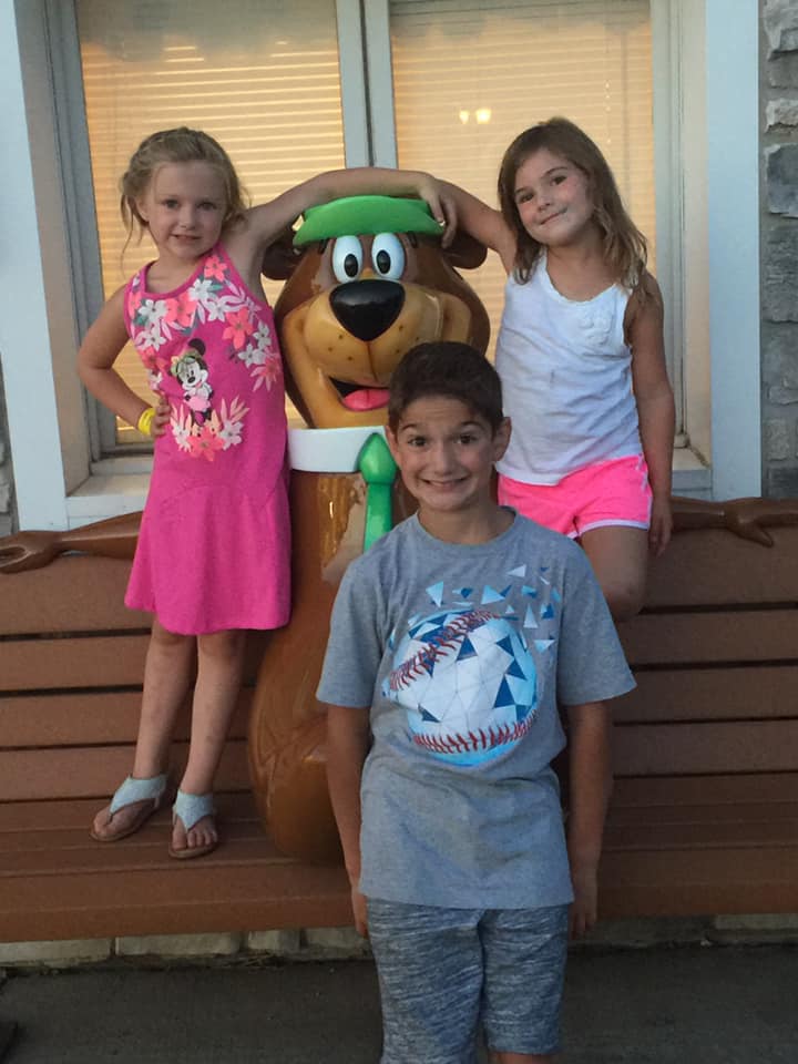 Two young girls stand on either side of a Yogi Bear statue on a bench, with a boy in front of them, all smiling at the camera.