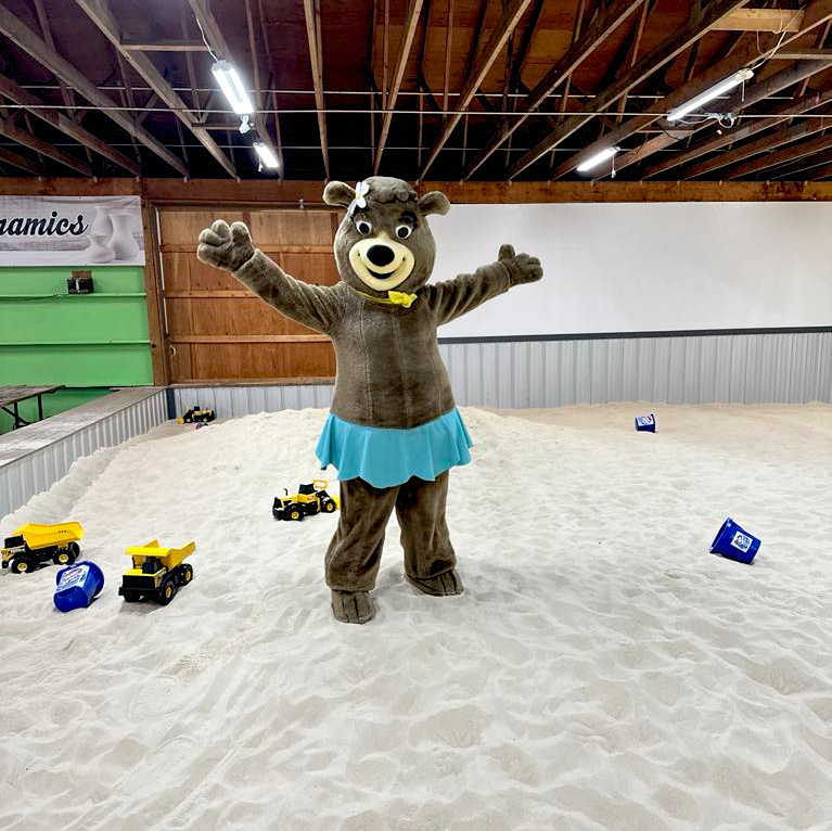 Person in a bear costume with a blue skirt stands with arms outstretched in an indoor sandbox with toy trucks and buckets scattered around.