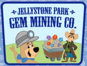 Cartoon sign reads "Jellystone Park Gem Mining Co." with a bear in mining gear and a man standing beside a cart filled with colorful gems.