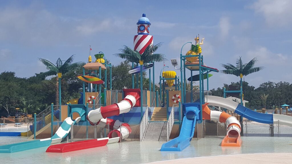 Colorful water playground with slides, palm tree decorations, a lighthouse structure, and shallow pool under a blue sky.